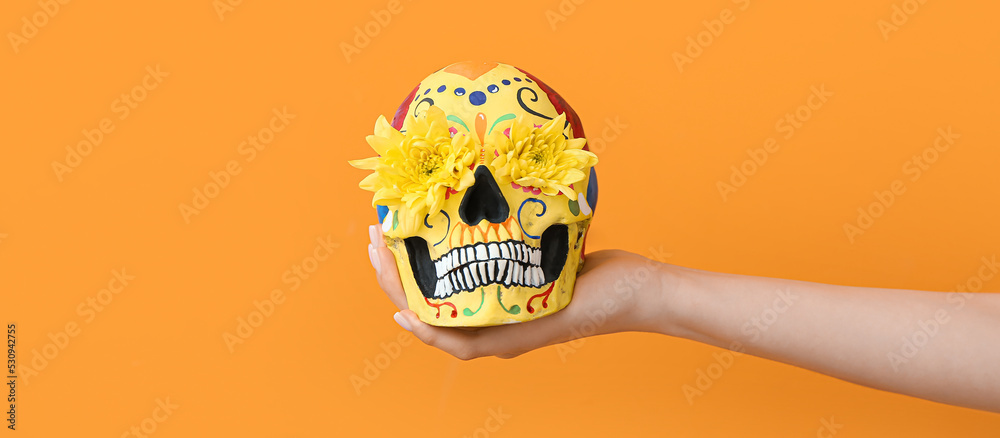 Hand holding painted skull and flowers on orange background. Celebration of Mexico's Day of the Dead (El Dia de Muertos) - obrazy, fototapety, plakaty 