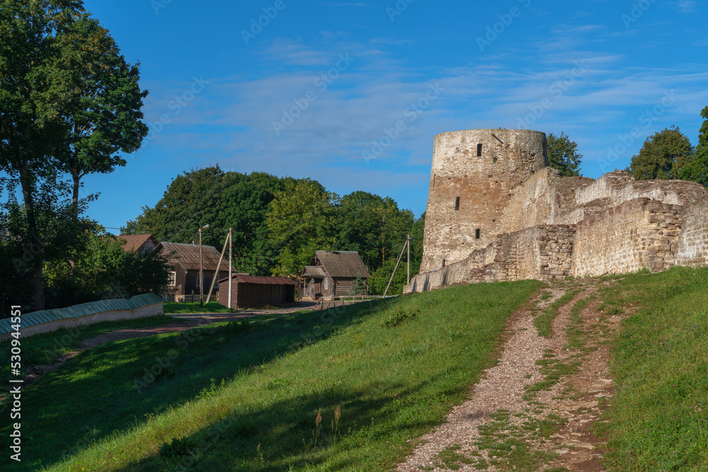 View of the wall and tower of the Izborsk fortress against the background of a village on a sunny summer day, Izborsk, Pskov region, Russia