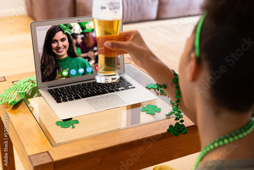 Mixed race woman holding beer having st patrick's day video call with friend on laptop at home