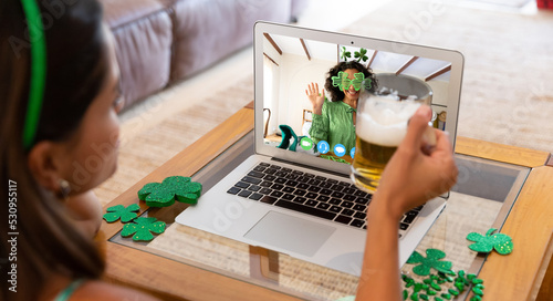 Caucasian woman holding beer having st patrick's day video call with friend on laptop at home
