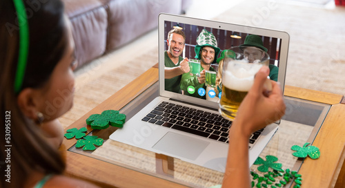 Caucasian woman holding beer having st patrick's day video call with friends on laptop at home