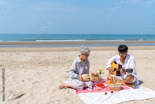 Happy Asian Family senior couple on summer beach vacation. Elderly retired man and woman enjoy and fun outdoor lifestyle playing guitar and singing together while travel and picnic at the beach.