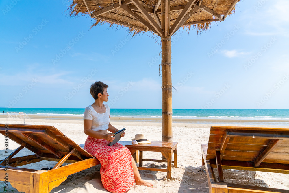 Modern Asian businesswoman working on digital tablet with internet at tropical beach in summer sunny day. Woman freelancer enjoy outdoor lifestyle work and travel on summer beach holiday vacation.