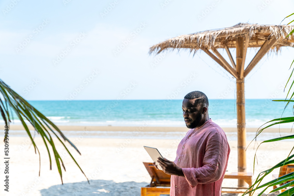 Modern African businessman working on digital tablet for business stocks trading on tropical beach in summer sunny day. Freelancer guy enjoy outdoor lifestyle work and travel on summer travel vacation