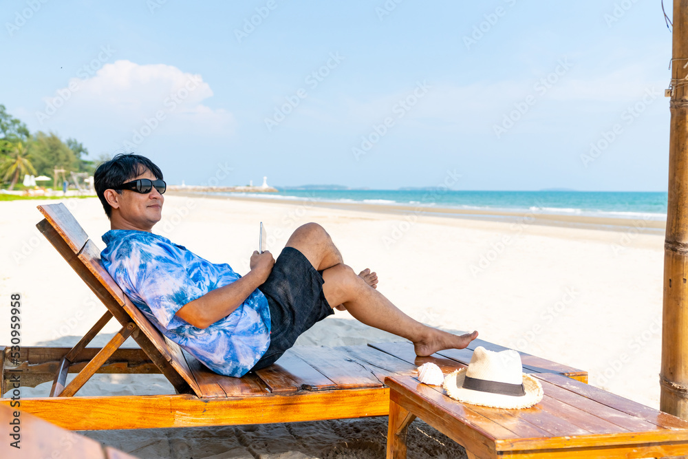 Modern Asian senior businessman working on digital tablet with internet at tropical beach in sunny day. Elderly retired man enjoy outdoor lifestyle with using technology on summer holiday vacation