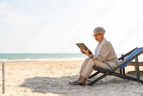 Modern Asian senior businesswoman working on digital tablet with internet at tropical beach in sunny day. Elderly retired woman enjoy outdoor lifestyle with using technology on summer holiday vacation