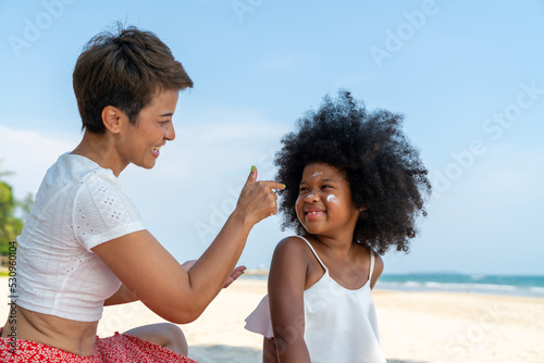 Happy African family on summer beach holiday vacation. Mother applying sunblock to little daughter. Mom and child girl kid enjoy outdoor lifestyle walking together on tropical beach at summer sunset.