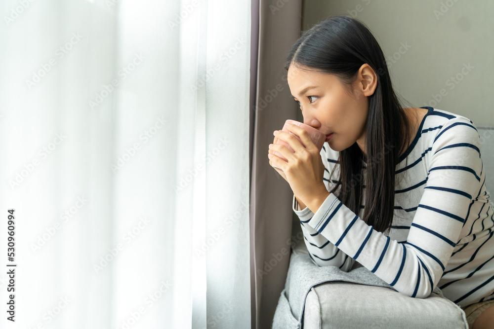 Young beautiful Asian woman standing by the window and looking to cityscape with drinking hot coffee or tea in the morning. Happy girl relax and enjoy indoors lifestyle on holiday vacation at home. Young beautiful Asian woman standing by the window and looking to cityscape with drinking hot coffee or tea in the morning. Happy girl relax and enjoy indoors lifestyle on holiday vacation at home.