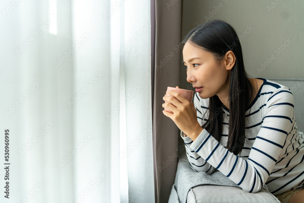 Young beautiful Asian woman standing by the window and looking to cityscape with drinking hot coffee or tea in the morning. Happy girl relax and enjoy indoors lifestyle on holiday vacation at home. Young beautiful Asian woman standing by the window and looking to cityscape with drinking hot coffee or tea in the morning. Happy girl relax and enjoy indoors lifestyle on holiday vacation at home.