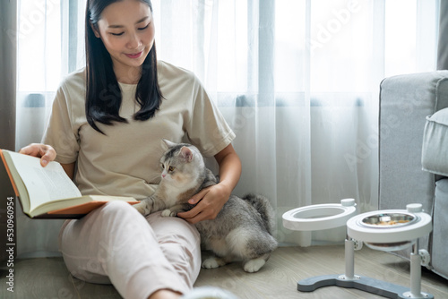 Young beautiful Asian woman relaxing with white tabby cat in apartment living room and reading a book. Happy girl relax and enjoy indoors lifestyle and leisure activity with pet on vacation at home.