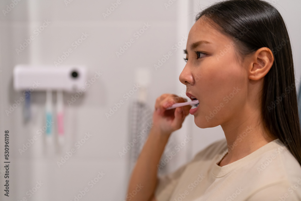 Smiling young attractive Asian woman brushing teeth and looking to the mirror in bathroom. Happy girl using toothbrush with whitening toothpaste cleaning teeth in the morning. Dental care concept. Smiling young attractive Asian woman brushing teeth and looking to the mirror in bathroom. Happy girl using toothbrush with whitening toothpaste cleaning teeth in the morning. Dental care concept.