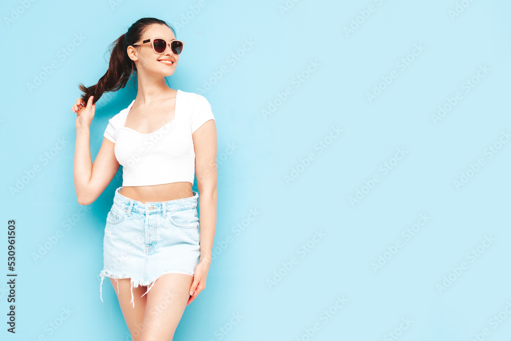 Young beautiful smiling female in trendy summer jeans skirt and top  clothes. Sexy carefree woman posing near blue wall in studio. Positive brunette model having fun.Cheerful and happy. In sunglasses