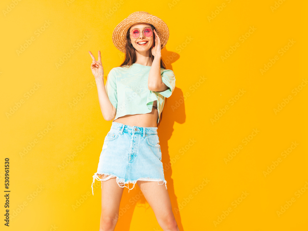 Portrait of young beautiful smiling female in trendy summer jeans skirt. carefree woman posing near yellow wall in studio. Positive model having fun indoors in sunglasses. Cheerful and happy. In hat Portrait of young beautiful smiling female in trendy summer jeans skirt. carefree woman posing near yellow wall in studio. Positive model having fun indoors in sunglasses. Cheerful and happy. In hat