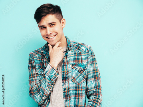 Portrait of handsome smiling model. Sexy stylish man dressed in checkered shirt and jeans. Fashion hipster male posing near blue wall in studio. Isolated. Touching his chin