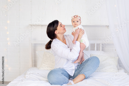 mom and baby girl hug and kiss sitting on a white cotton bed at home, maternal love and care