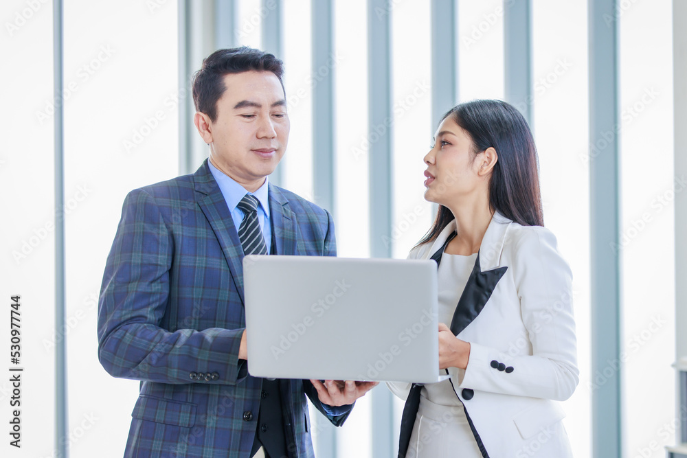 Millennial Asian successful professional male businessman manager in formal suit standing smiling holding laptop computer talking helping advising female businesswoman colleague in company office Millennial Asian successful professional male businessman manager in formal suit standing smiling holding laptop computer talking helping advising female businesswoman colleague in company office