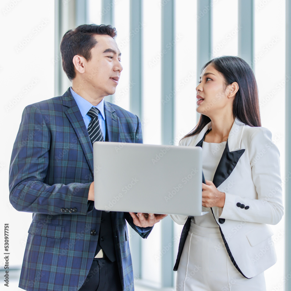 Millennial Asian successful professional male businessman manager in formal suit standing smiling holding laptop computer talking helping advising female businesswoman colleague in company office