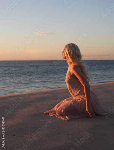 full length portrait of beautiful young woman with long hair wearing flowing dress  sitting pose at ocean beach background with sunset lighting.