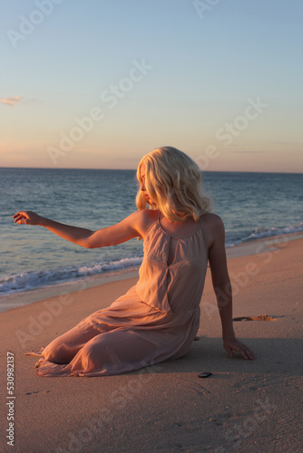 full length portrait of beautiful young woman with long hair wearing flowing dress, sitting pose at ocean beach background with sunset lighting.