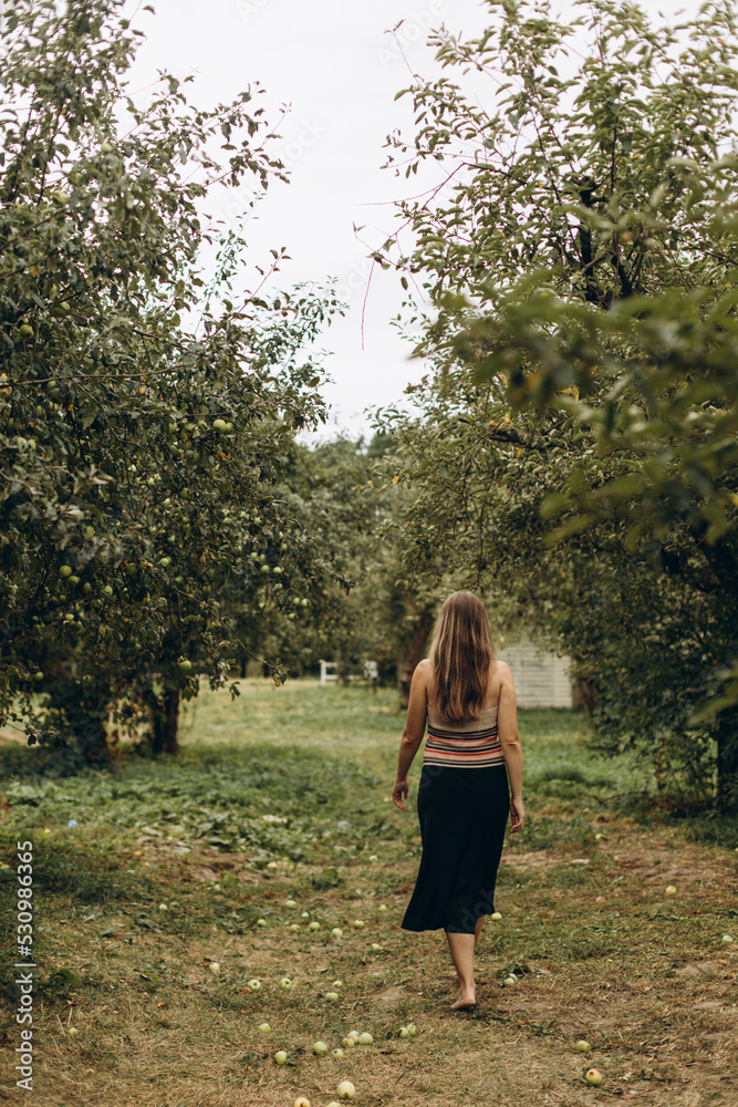 a woman in a black skirt walks barefoot on the trampled grass in an apple orchard. a girl with streaked long hair walks alone in her garden on a cloudy day. a woman in a black skirt walks barefoot on the trampled grass in an apple orchard. a girl with streaked long hair walks alone in her garden on a cloudy day.