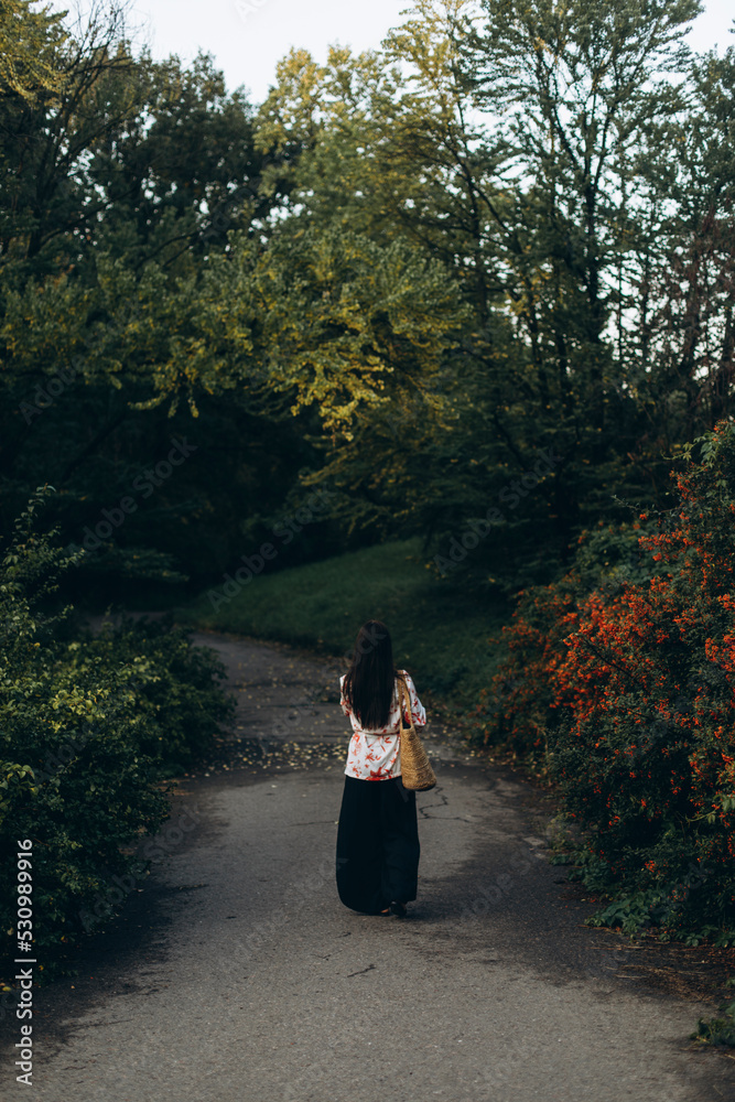 young beautiful woman walking in the park rear view. a stylish girl in black wide trousers and a straw bag walks through the city park. young beautiful woman walking in the park rear view. a stylish girl in black wide trousers and a straw bag walks through the city park.