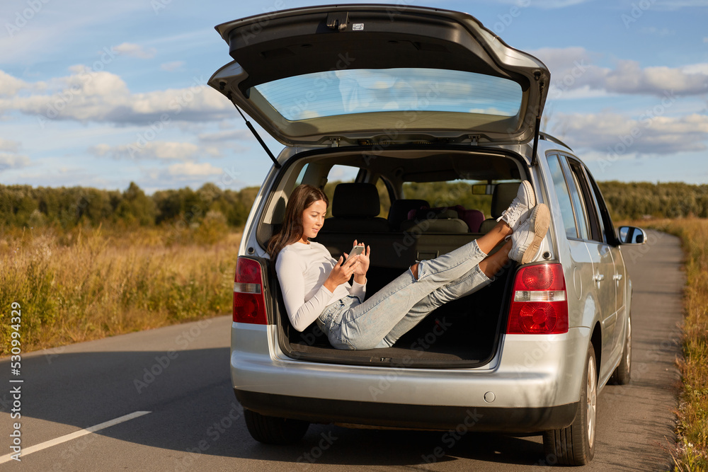 Image of dark haired Caucasian young adult woman sitting in car trunk, female wearing white shirt and jeans, resting while traveling and using cell phone, enjoying good weather and her journey.