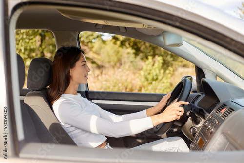 Profile portrait of delighted satisfied beautiful female with glad positive expression, being satisfied with riding car alone, sits on driver`s seat, looking at road with positive expression.