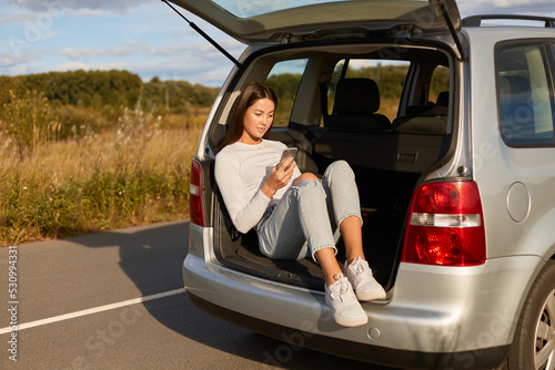 Image of dark haired young adult woman traveler sitting in car trunk and using cell phone, female wearing casual clothes, surfing internet, checking social networks while traveling.