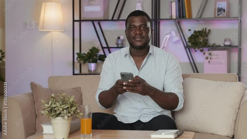 Medium portrait of African American man posing for camera smiling and holding smartphone in hands, sitting on beige sofa in contemporary minimalist style apartment Medium portrait of African American man posing for camera smiling and holding smartphone in hands, sitting on beige sofa in contemporary minimalist style apartment
