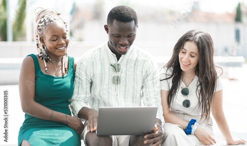 Gruppo ridente di giovani multietnici d'affari che lavorano su un laptop seduti insieme su una panchina in centro cita photo
