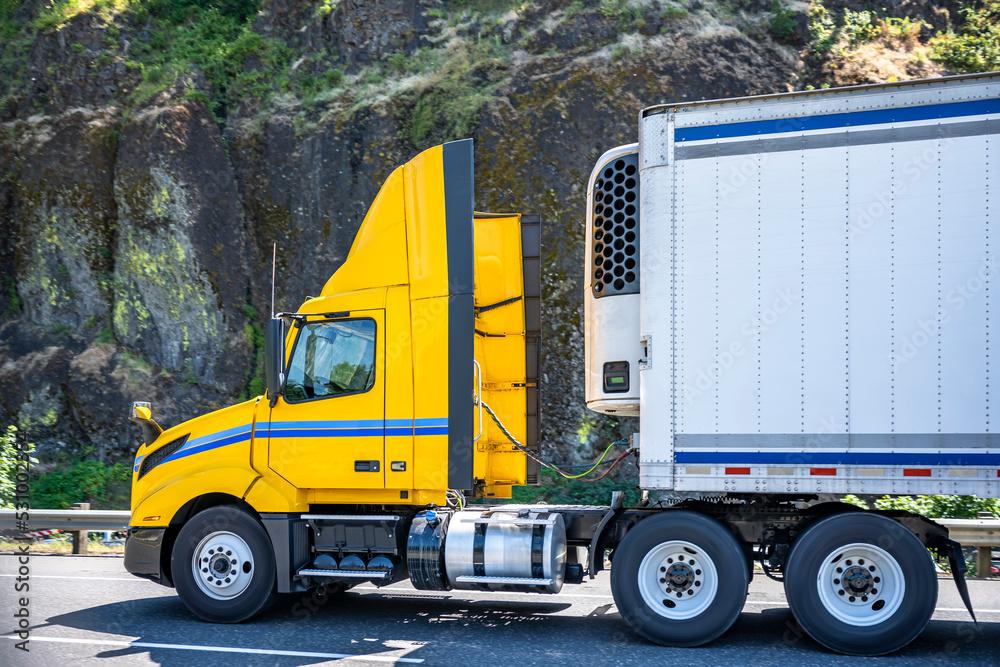 Bright yellow big rig day cab semi truck with roof spoiler transporting food cargo in reefer semi trailer running on the mountain road with rock wall on the side Bright yellow big rig day cab semi truck with roof spoiler transporting food cargo in reefer semi trailer running on the mountain road with rock wall on the side