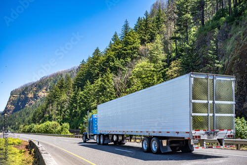 Classic blue American bonnet big rig semi truck transporting cargo in refrigerator semi trailer driving on the divided highway road with mountain and wild forest