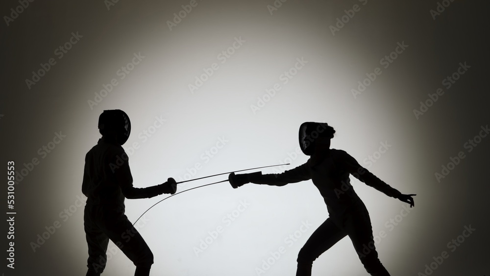 Side view of black silhouettes two young female swordswoman with crossed rapiers. Athletes in white uniforms and protective helmet fighting a duel in dark studio with backlight.