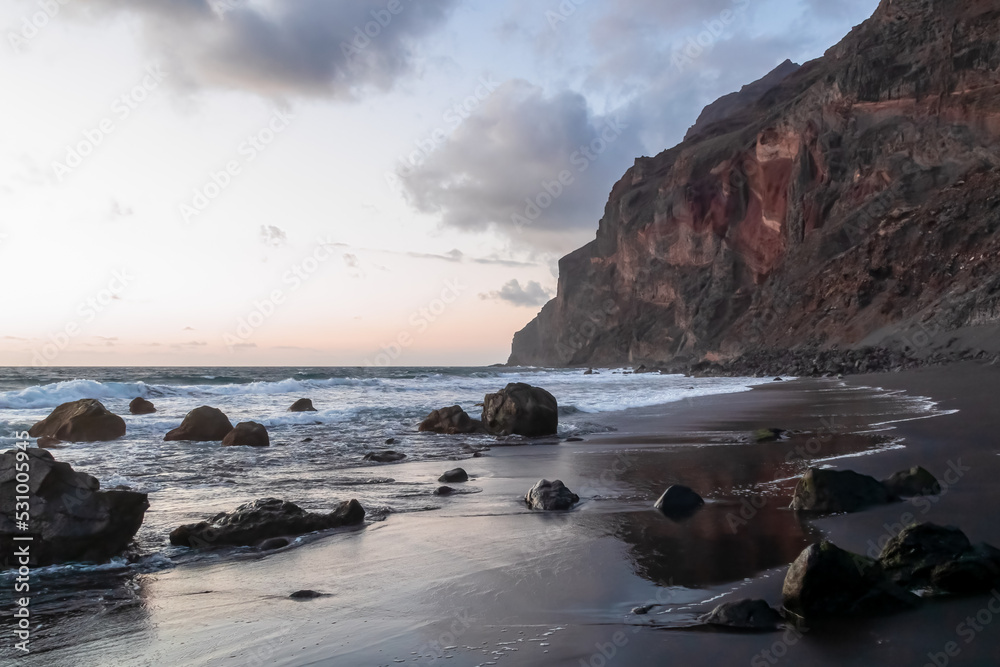 Scenic view during sunset on volcanic black sand beach Playa del Ingles in Valle Gran Rey, La Gomera, Canary Islands, Spain, Europe. Massive cliffs of the La Mercia range. Smooth waves sweeping rocks Scenic view during sunset on volcanic black sand beach Playa del Ingles in Valle Gran Rey, La Gomera, Canary Islands, Spain, Europe. Massive cliffs of the La Mercia range. Smooth waves sweeping rocks