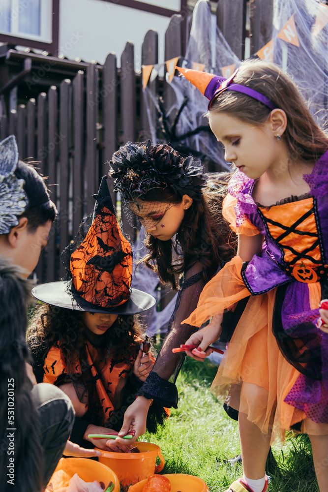 Multiethnic kids in costumes taking candies from buckets during halloween celebration outdoors Multiethnic kids in costumes taking candies from buckets during halloween celebration outdoors