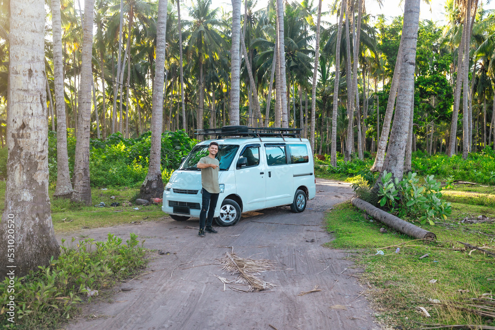 young man in front of white camper van parked in tropical scenic field of Bali Indonesia at sunset