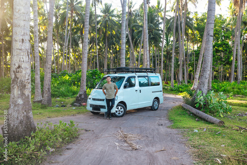 happy young man standing in front of white camper van parked on dirt road in tropical coconut tree field in Bali Indonesia at sunset