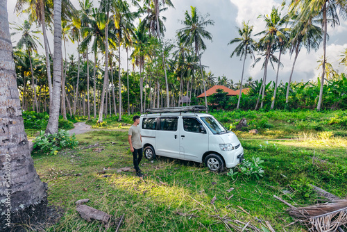young man with beautiful camper van parked in a tropical scenic field with coconut trees in Bali Indonesia at sunset