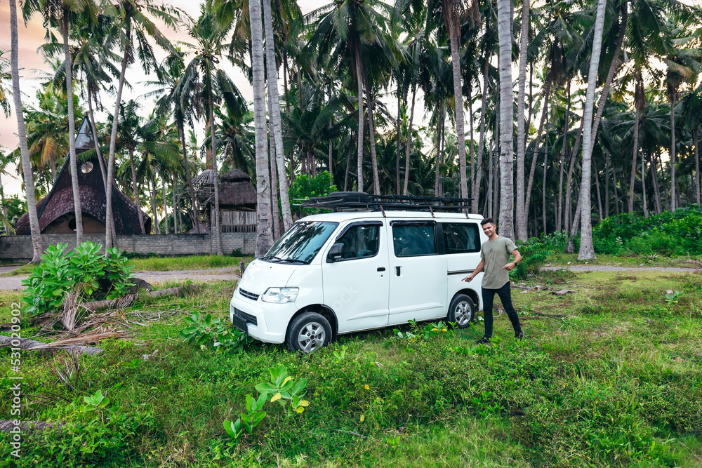 wide landscape of young man standing next to white camper van parked in tropical field with coconut trees at sunset in Bali Indonesia
