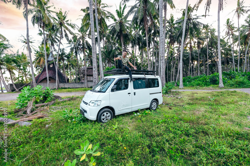 young happy tourist sitting on top of white camper van parked in a tropical coconut tree field in Bali Indonesia young happy tourist sitting on top of white camper van parked in a tropical coconut tree field in Bali Indonesia
