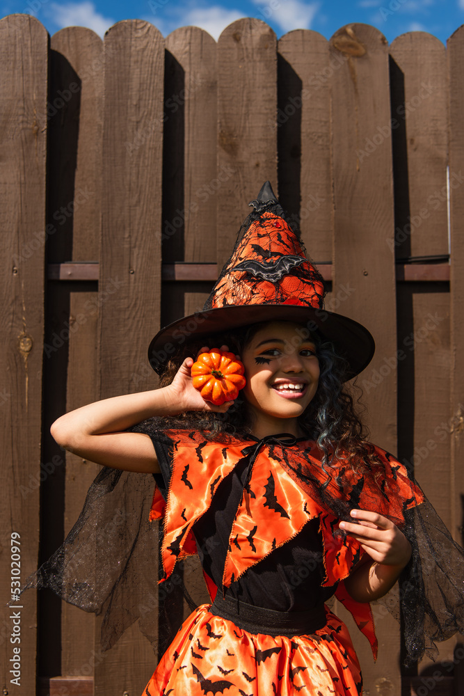cheerful curly girl in halloween costume and witch hat holding small pumpkin and looking at camera