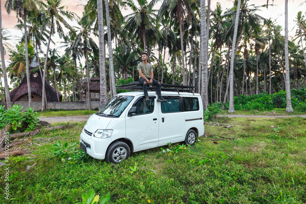 young man sitting on top of white camper van parked in a tropical green coconut tree field in Bali Indonesia