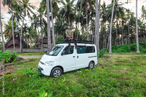 young man sitting on top of white camper van parked in a tropical green coconut tree field in Bali Indonesia