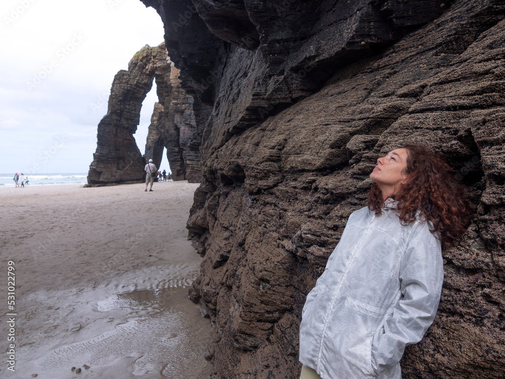 Brunette female with white rain coat looking at the sky with dreamy look leaning against  magical arch rock formations.