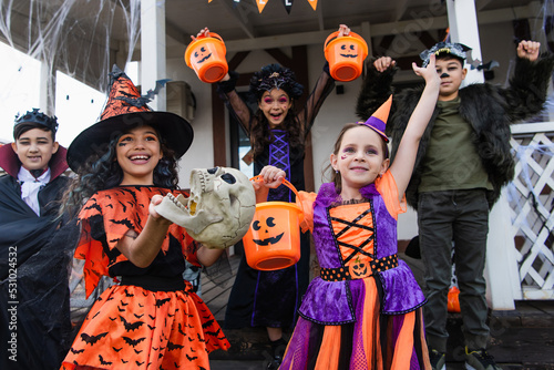 excited multicultural friends in halloween costumes holding trick or treat buckets near house
