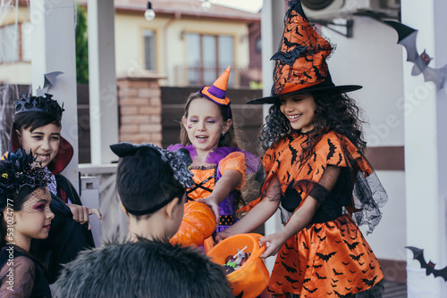 Smiling kid in halloween costume holding bucket with candies near interracial friends outdoors