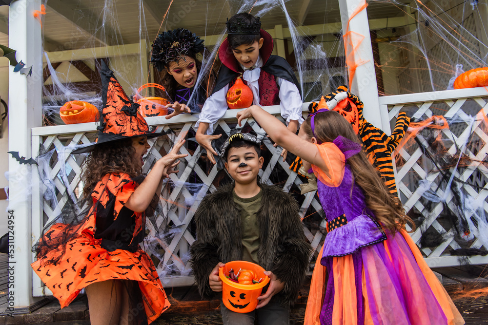 Preteen children in halloween costumes grimacing near asian friend with bucket outdoors
