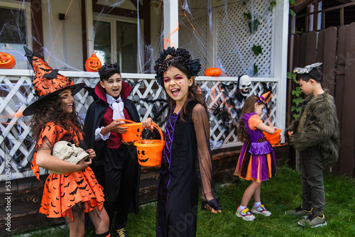 Cheerful girl holding halloween bucket near interracial friends with skull outdoors