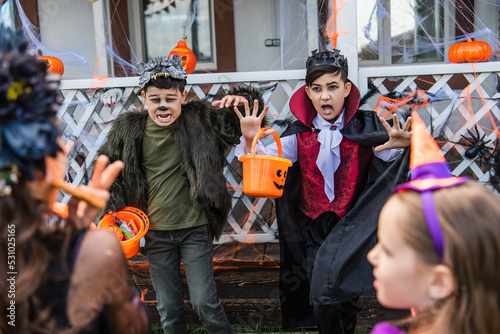 Asian kids in halloween costumes holding buckets with candies near blurred friends in backyard