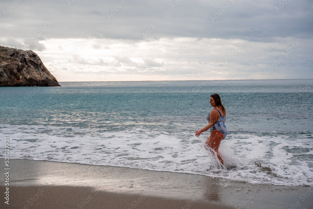 A plump woman in a bathing suit enters the water during the surf. Alone on the beach, Gray sky in the clouds, swimming in winter. A plump woman in a bathing suit enters the water during the surf. Alone on the beach, Gray sky in the clouds, swimming in winter.